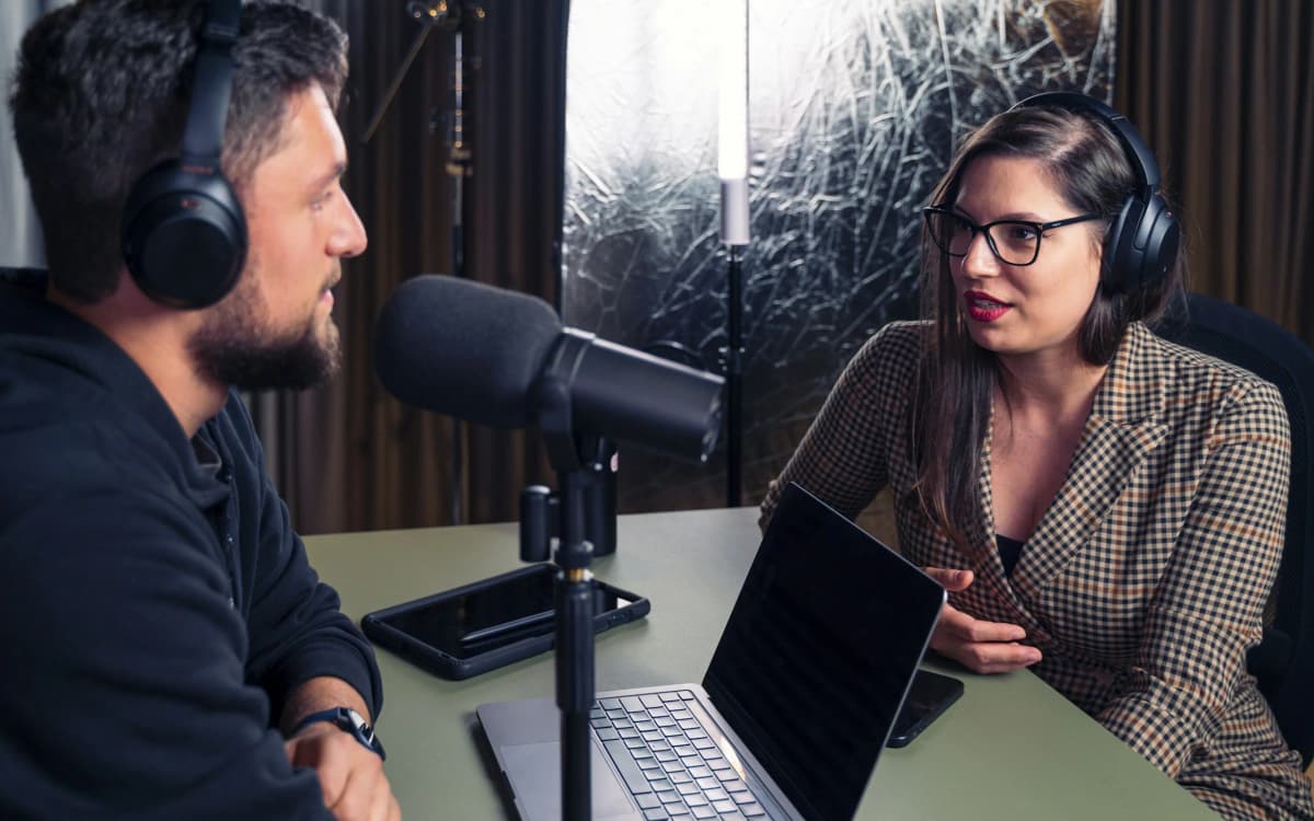 Two people in headphones talking across a table during a podcast recording